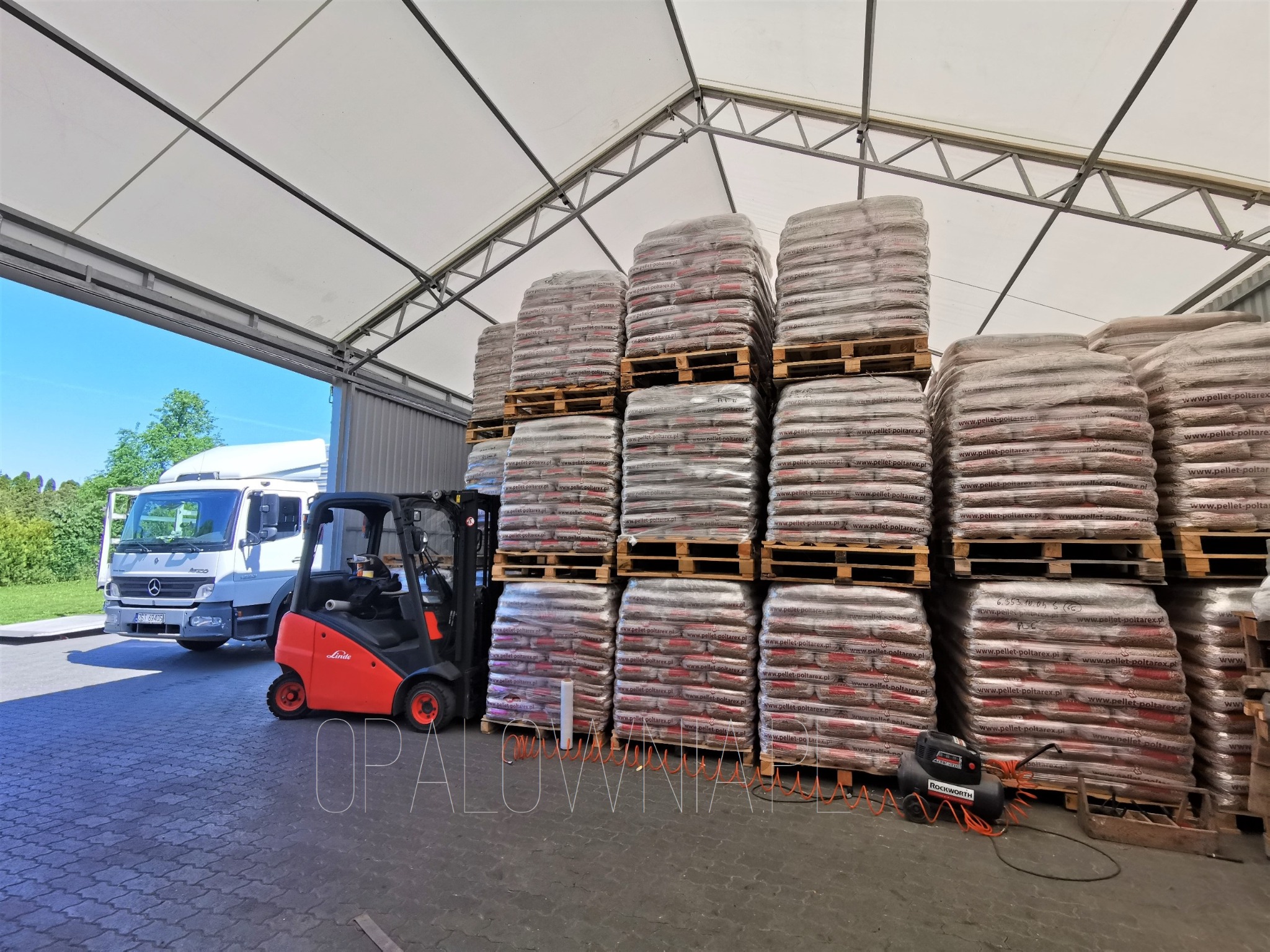 Magazyn pelletu: wózek widłowy Linde obok palet z pelletem poltarex.pl pod zadaszeniem, w tle biała ciężarówka Mercedes
