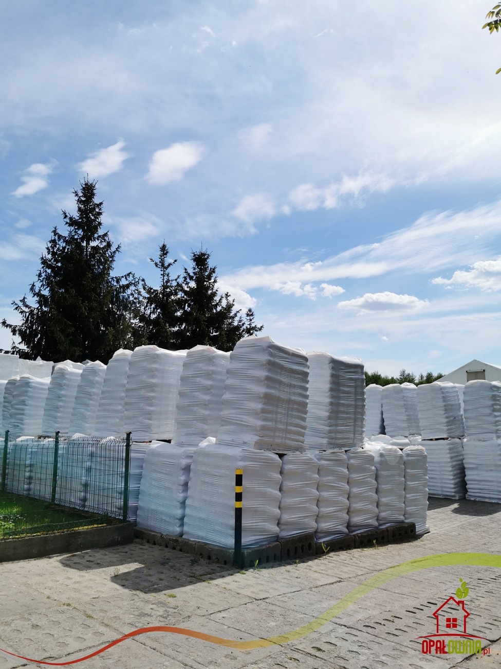 Składowisko workowanego ekogroszku na paletach, zabezpieczonego folią, na betonowym placu pod błękitnym niebem z widocznymi drzewami iglastymi w tle.