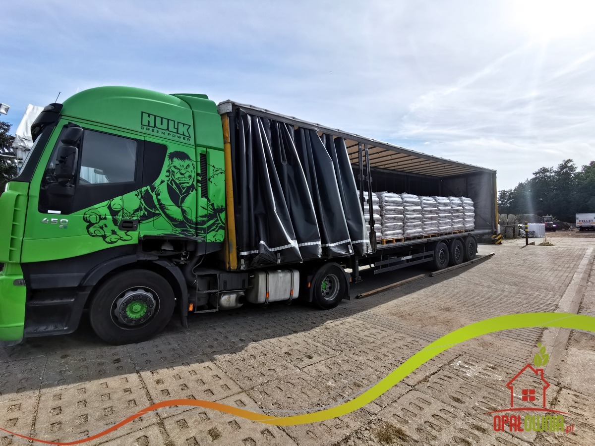 Zielona ciężarówka z grafiką Hulka przewożąca palety z workami, częściowo odsłonięta plandeka, kostka brukowa na placu.