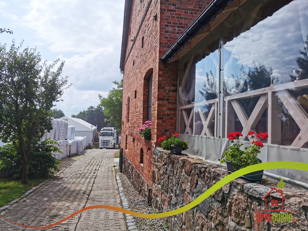 Ceglany budynek z przeszkloną werandą, kwiaty w donicach na kamiennym murku, w tle biały namiot i ciężarówka z towarem na brukowanej drodze.
