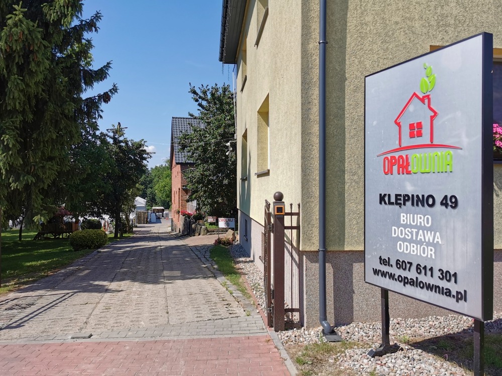 Tablica informacyjna z logo 'Opałownia' i adresem Klepino 49, widoczna z chodnika prowadzącego do budynku firmy, z drzewami i fragmentem drugiego budynku w tle.