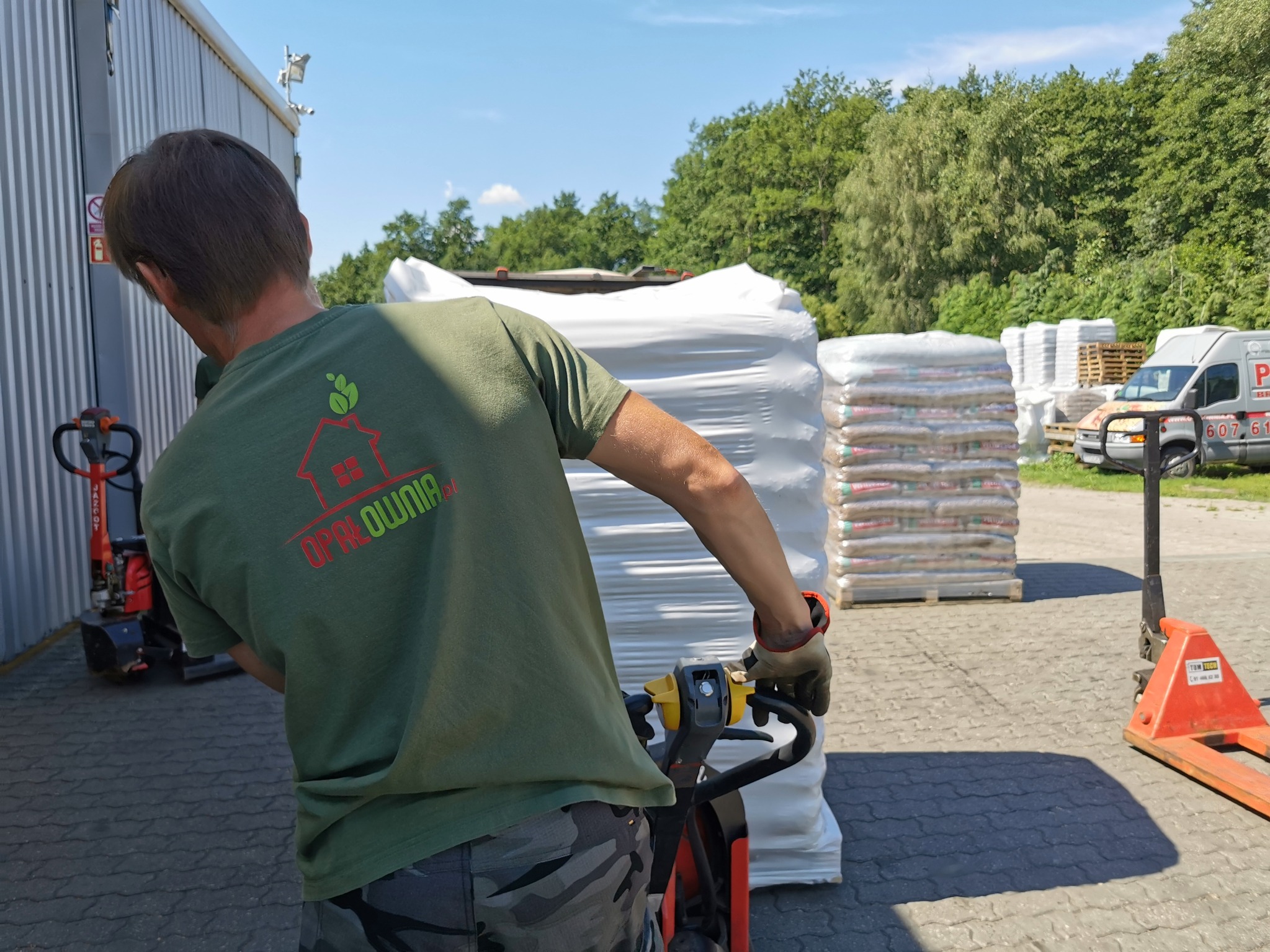 Mężczyzna w koszulce z logo firmy 'OPALOWNIA' obsługuje wózek paletowy z zapakowanymi workami, w tle skład opału i samochód dostawczy.