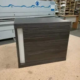 Ciemna szafka z szufladami o drewnopodobnej fakturze, z białą wstawką po lewej stronie, na tle maszyny stolarskiej w warsztacie.