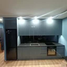 Nowoczesna kuchnia z szarymi, lakierowanymi frontami szafek, czarnym blatem i sprzętem AGD w zabudowie, oświetlona punktowymi lampami LED pod szafkami wiszącymi.