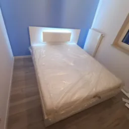 Białe łóżko z podświetlanym zagłówkiem, materac owinięty folią ochronną, ustawione w pokoju z niebieską ścianą.