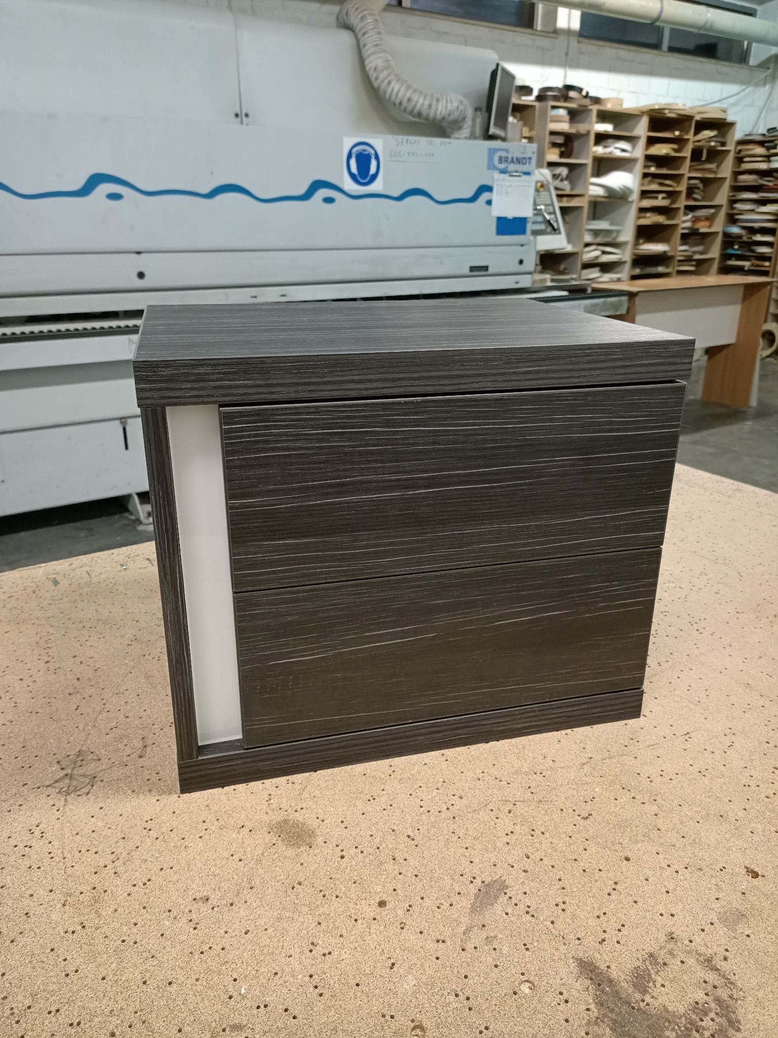 Ciemna szafka z szufladami o drewnopodobnej fakturze, z białą wstawką po lewej stronie, na tle maszyny stolarskiej w warsztacie.