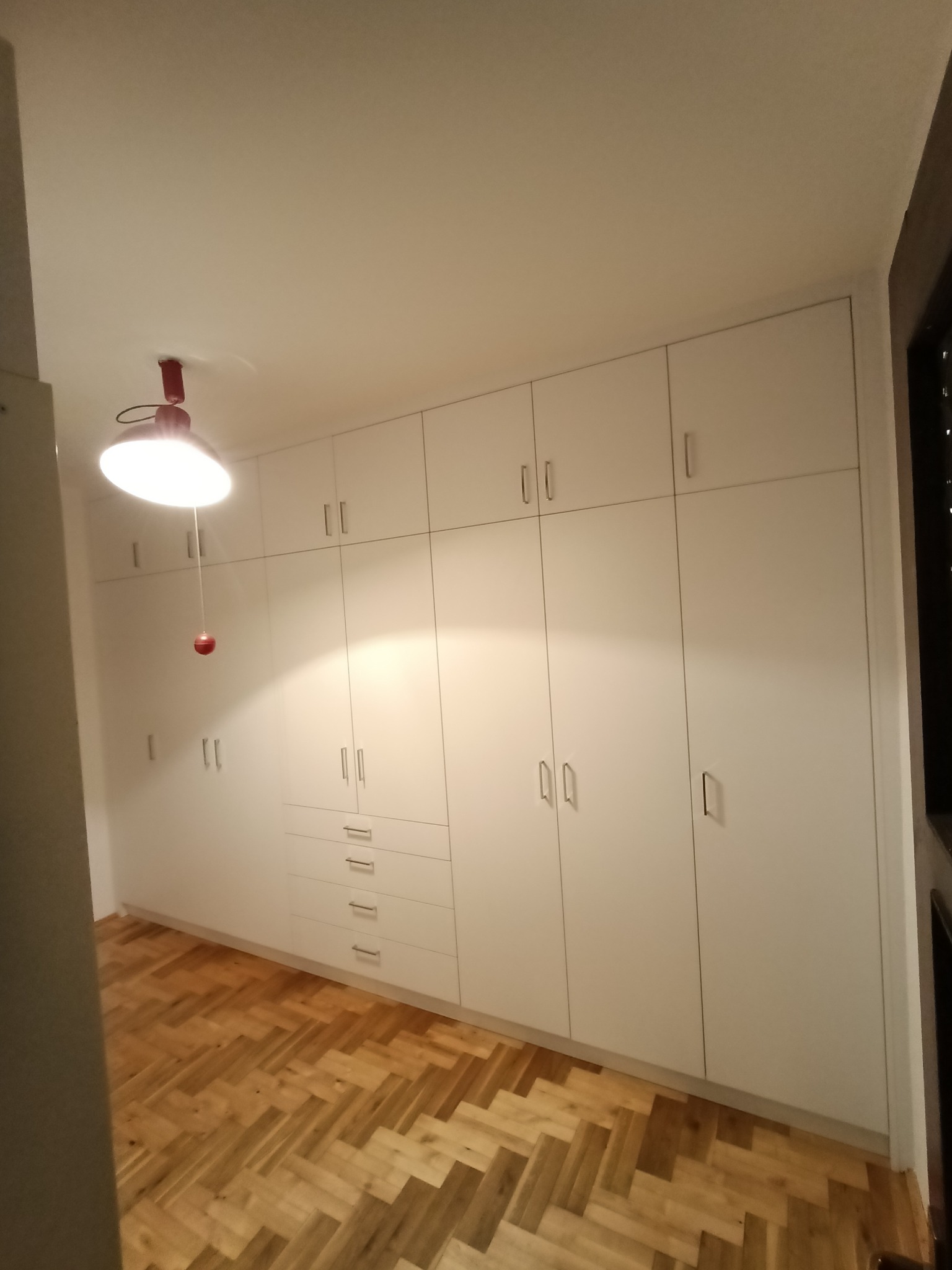 Biała zabudowa szaf wnękowych z szufladami i górnymi szafkami, parkiet w jodełkę na podłodze, oświetlenie sufitowe z czerwonym akcentem.