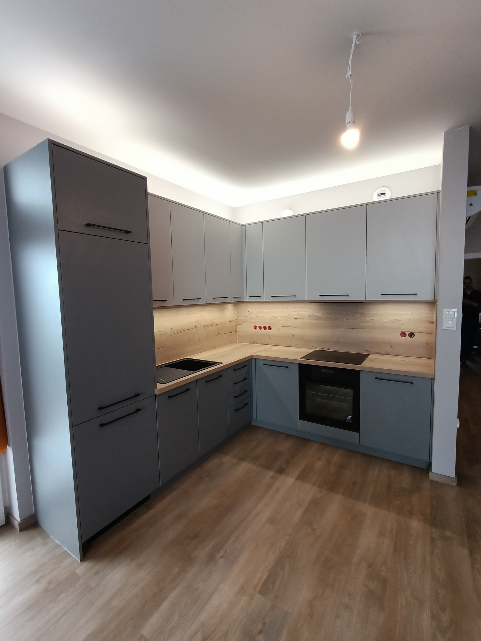 Nowoczesna kuchnia w odcieniach szarości z drewnianym blatem i oświetleniem LED pod szafkami, widok na narożnik z piekarnikiem i płytą indukcyjną.