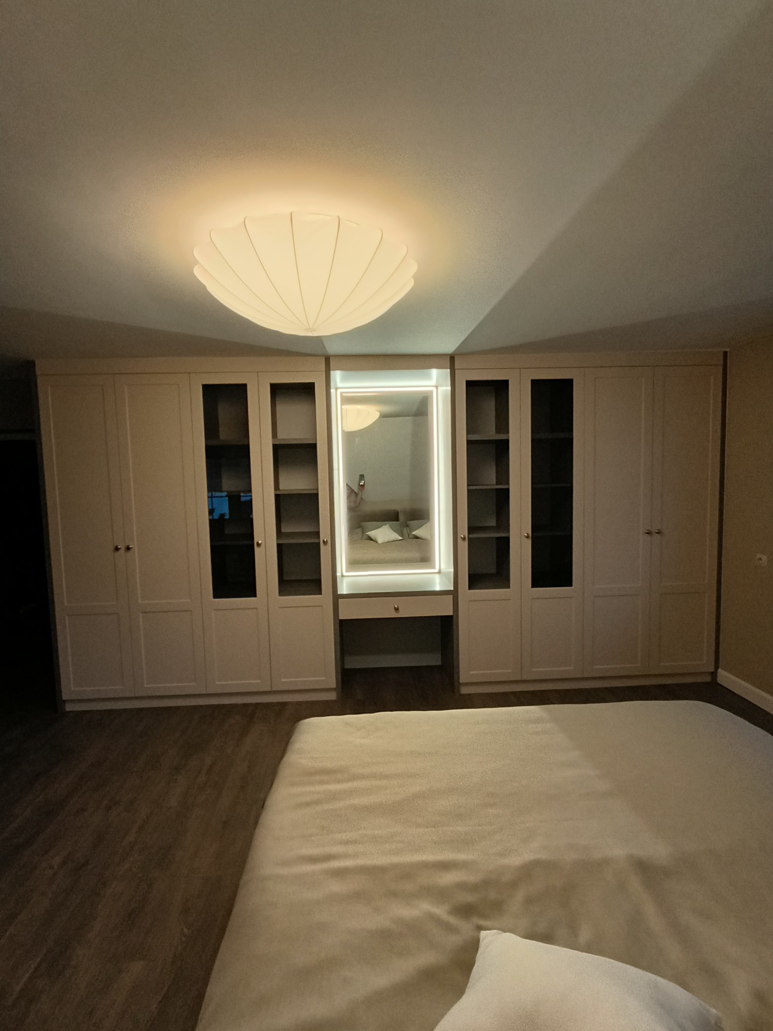 Jasna zabudowa szafy z przeszklonymi witrynami i toaletką z podświetlanym lustrem, widok z perspektywy łóżka w sypialni.