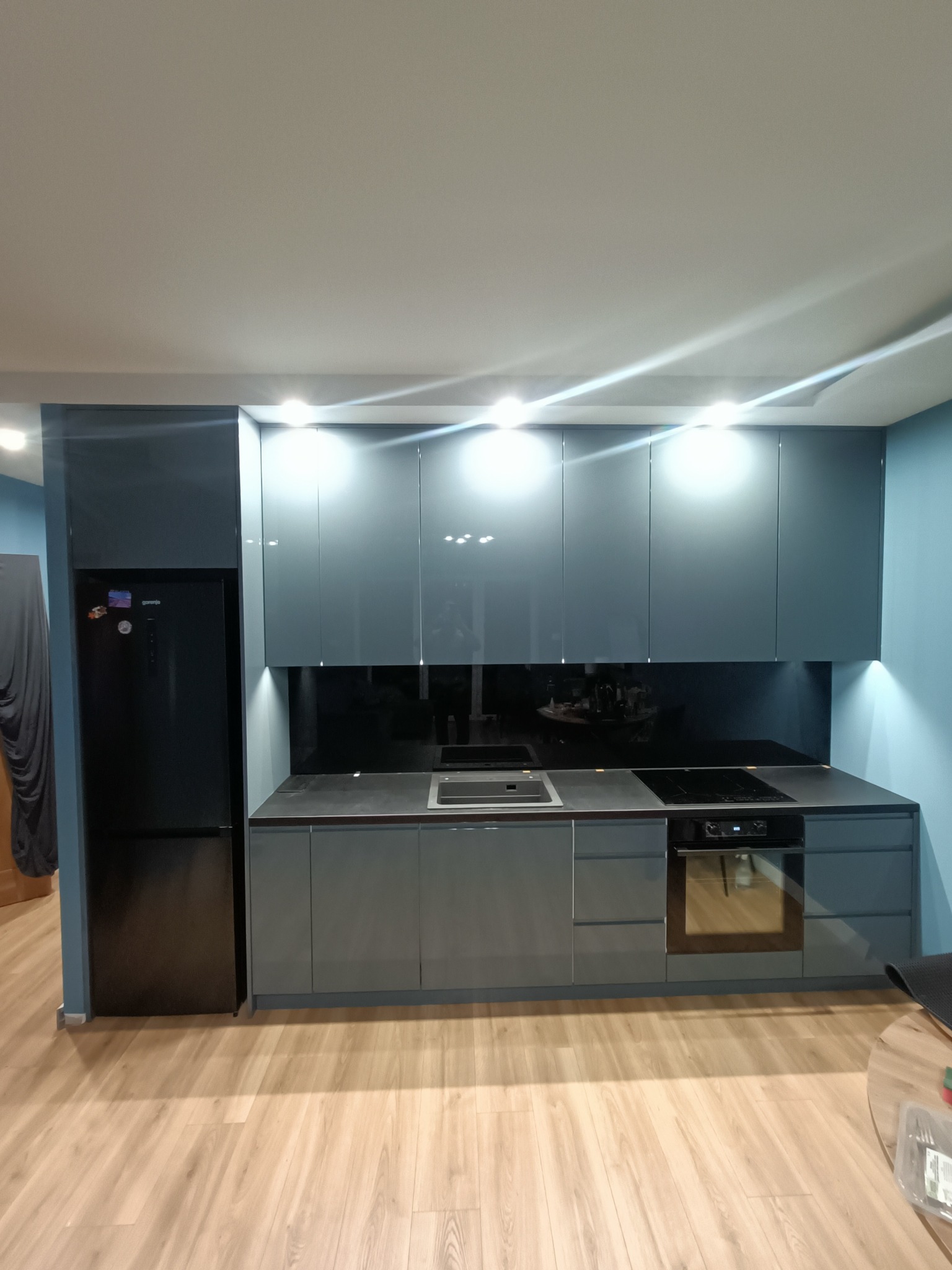 Nowoczesna kuchnia z szarymi, lakierowanymi frontami szafek, czarnym blatem i sprzętem AGD w zabudowie, oświetlona punktowymi lampami LED pod szafkami wiszącymi.