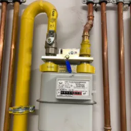 Zamontowany gazomierz Intergaz z żółtą rurą gazową i miedzianymi rurami instalacji grzewczej, widoczne mocowania rur do ściany.