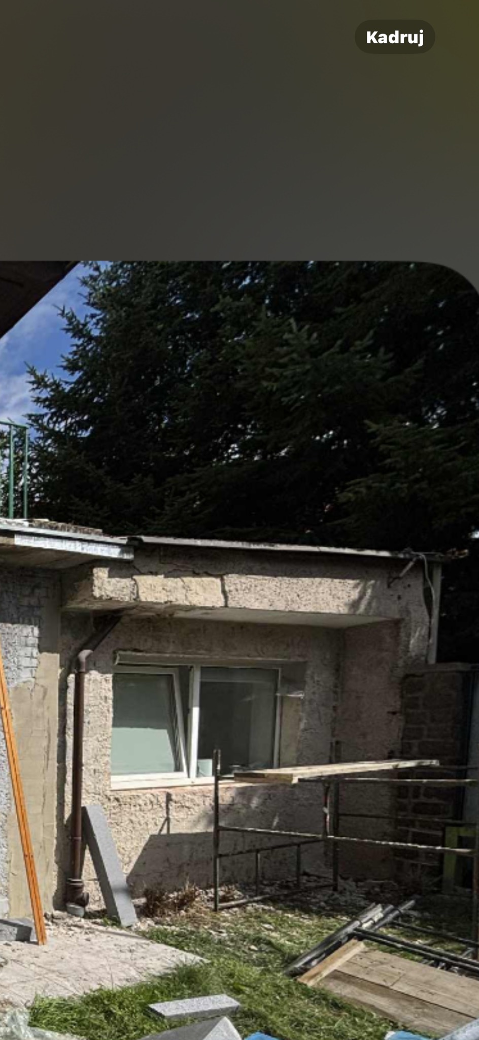 Niski budynek z oknem w trakcie prac elewacyjnych: widoczne rusztowanie, przygotowane płyty i fragmenty elewacji w stanie surowym. Tło: drzewa i fragment nieba.