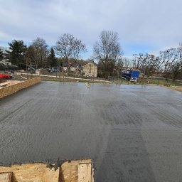 Wymurowany Sp. z o.o. - Wylewanie betonu na przygotowany fundament z drewnianym szalunkiem, widoczny pracownik w czerwonej bluzie, w tle budynki mieszkalne i błękitny samochód dostawczy.