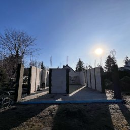 Wymurowany Sp. z o.o. - Wznoszenie ścian parteru domu z betonu komórkowego, widoczne zbrojenia pionowe, w tle drzewa i błękitne niebo z promieniami słońca.