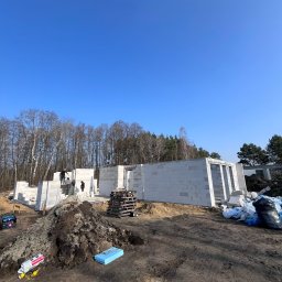 Wymurowany Sp. z o.o. - Budowa domu z betonu komórkowego w stanie surowym otwartym, widoczne palety, worki z materiałami budowlanymi i kopiec ziemi na pierwszym planie, dwóch pracowników na ścianach budynku.