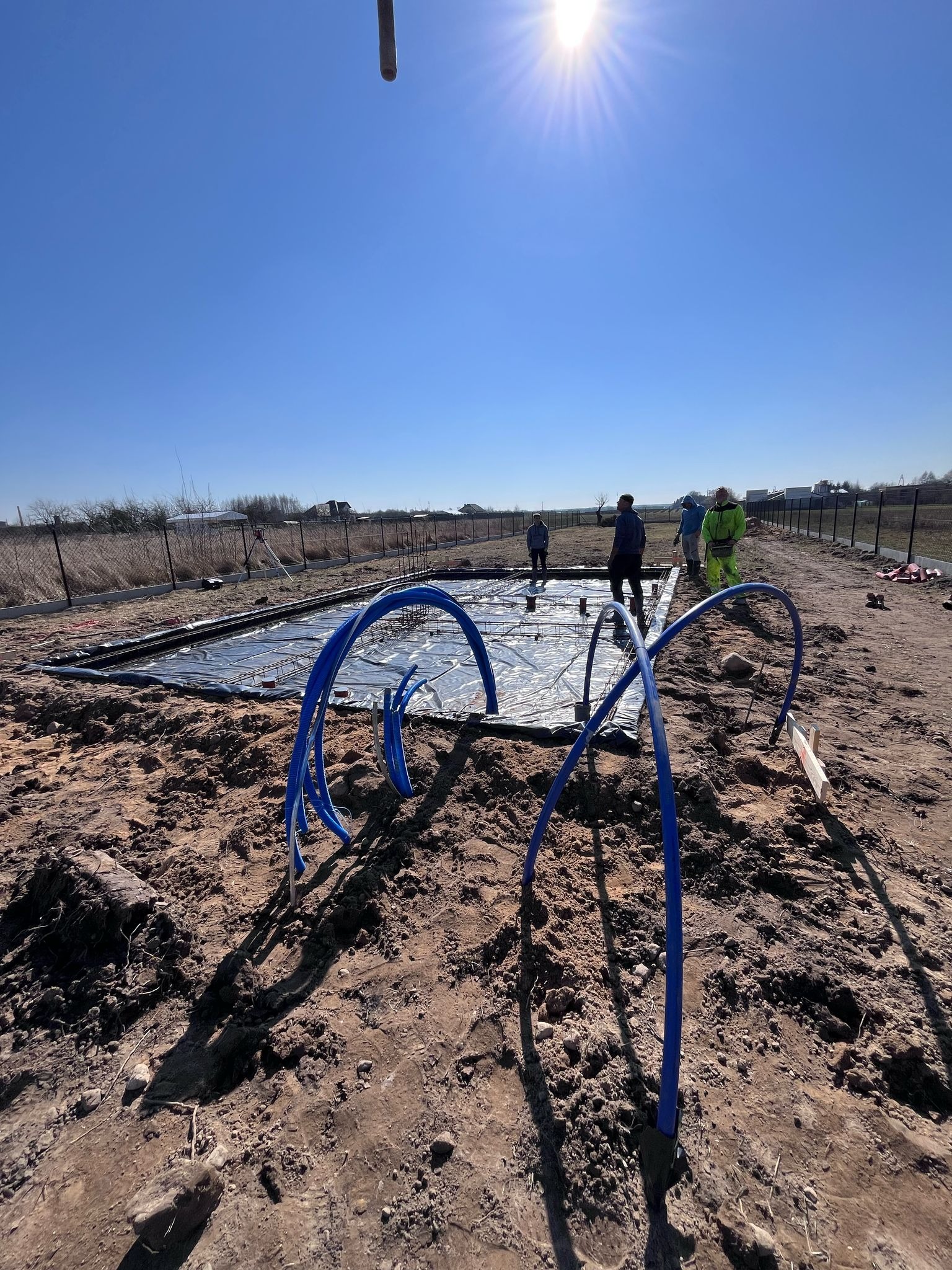 Przygotowanie terenu pod fundamenty z widoczną folią izolacyjną, zbrojeniem i niebieskimi rurami instalacyjnymi, ekipa budowlana w tle.