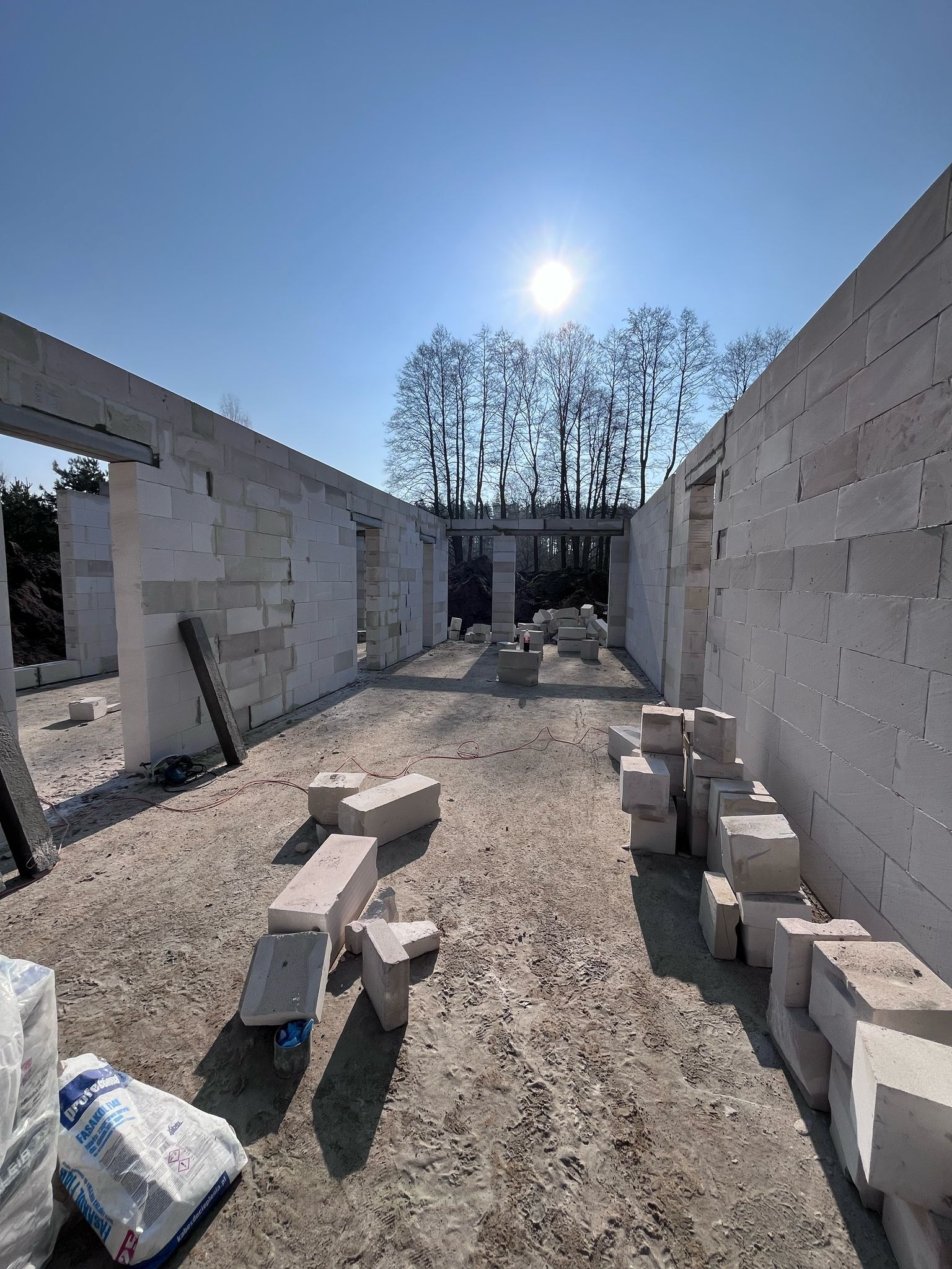 Wnętrze domu w trakcie budowy z białych bloczków, widoczne ściany, otwory okienne i drzwiowe, na podłodze porozrzucane bloczki i worki z zaprawą, w tle drzewa bez liści pod błękitnym niebem.