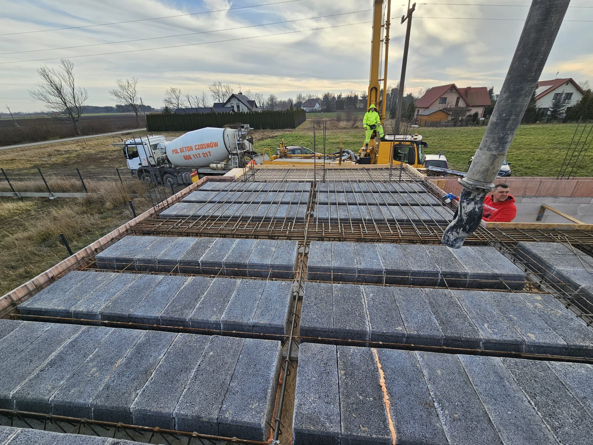 Wylewanie betonu na strop Teriva; widoczna pompa do betonu, pracownik w czerwonej bluzie i betoniarka na tle zabudowy jednorodzinnej.