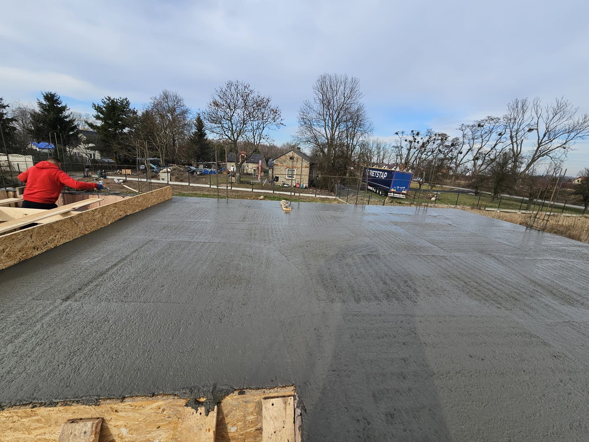Wylewanie betonu na przygotowany fundament z drewnianym szalunkiem, widoczny pracownik w czerwonej bluzie, w tle budynki mieszkalne i błękitny samochód dostawczy.