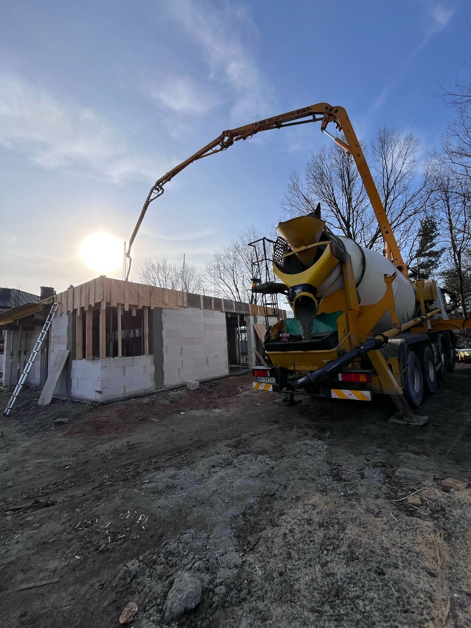 Wylewanie betonu z pompy do fundamentów domu w trakcie budowy, widoczna konstrukcja ścian z bloczków betonowych i drewniany szalunek stropu, na pierwszym planie pompa do betonu na podwoziu ciężarowym.