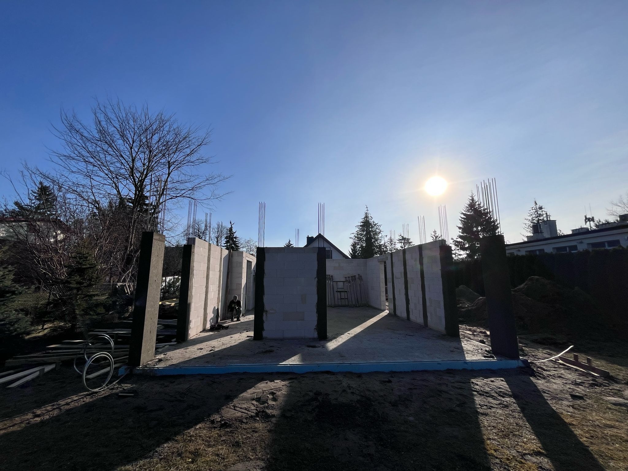 Wznoszenie ścian parteru domu z betonu komórkowego, widoczne zbrojenia pionowe, w tle drzewa i błękitne niebo z promieniami słońca.