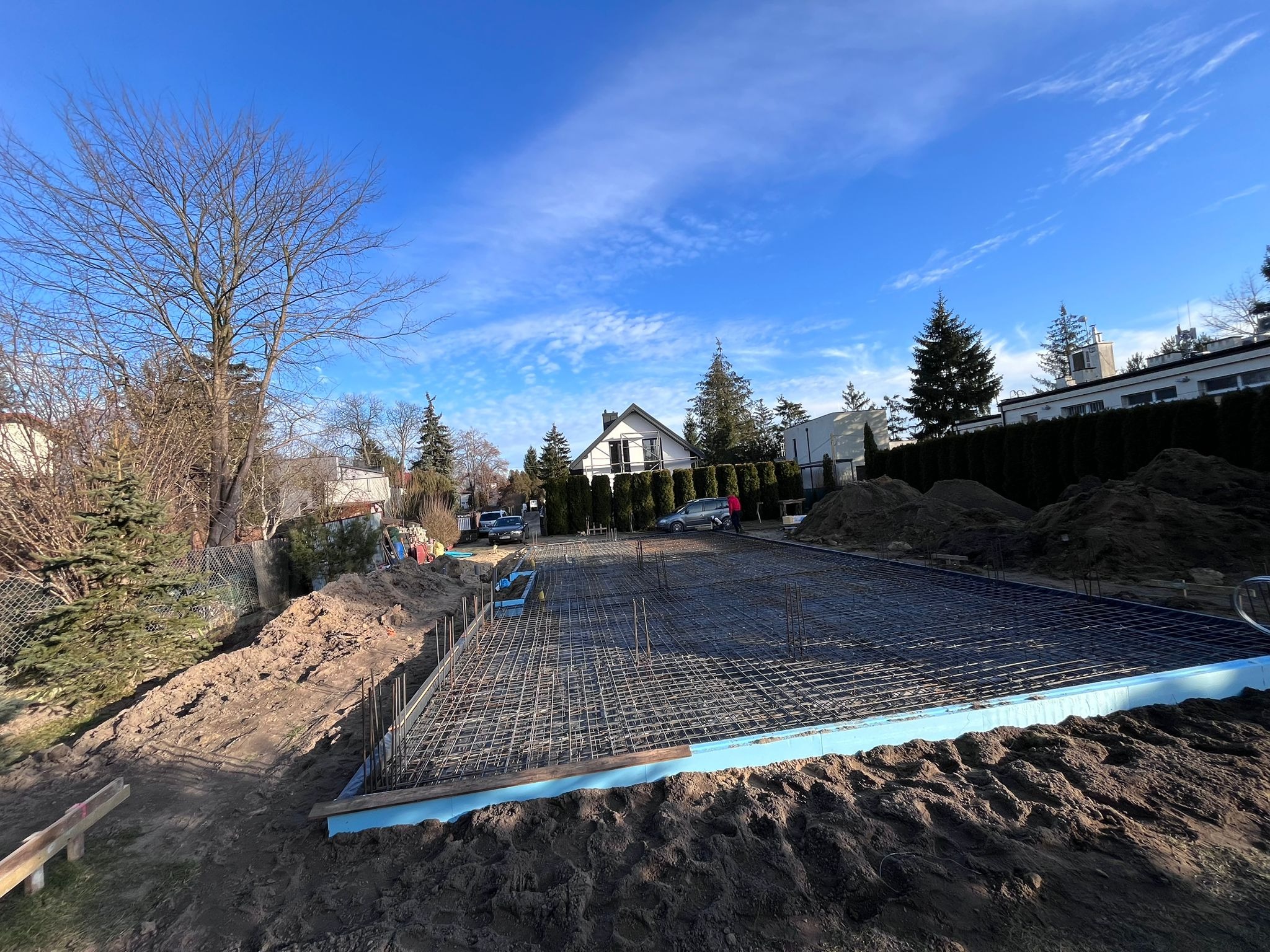 Zbrojenie fundamentu płyty, stalowa siatka na niebieskim styropianie, widok z góry na plac budowy z ziemią, drzewami i domami w tle, błękitne niebo z chmurami.