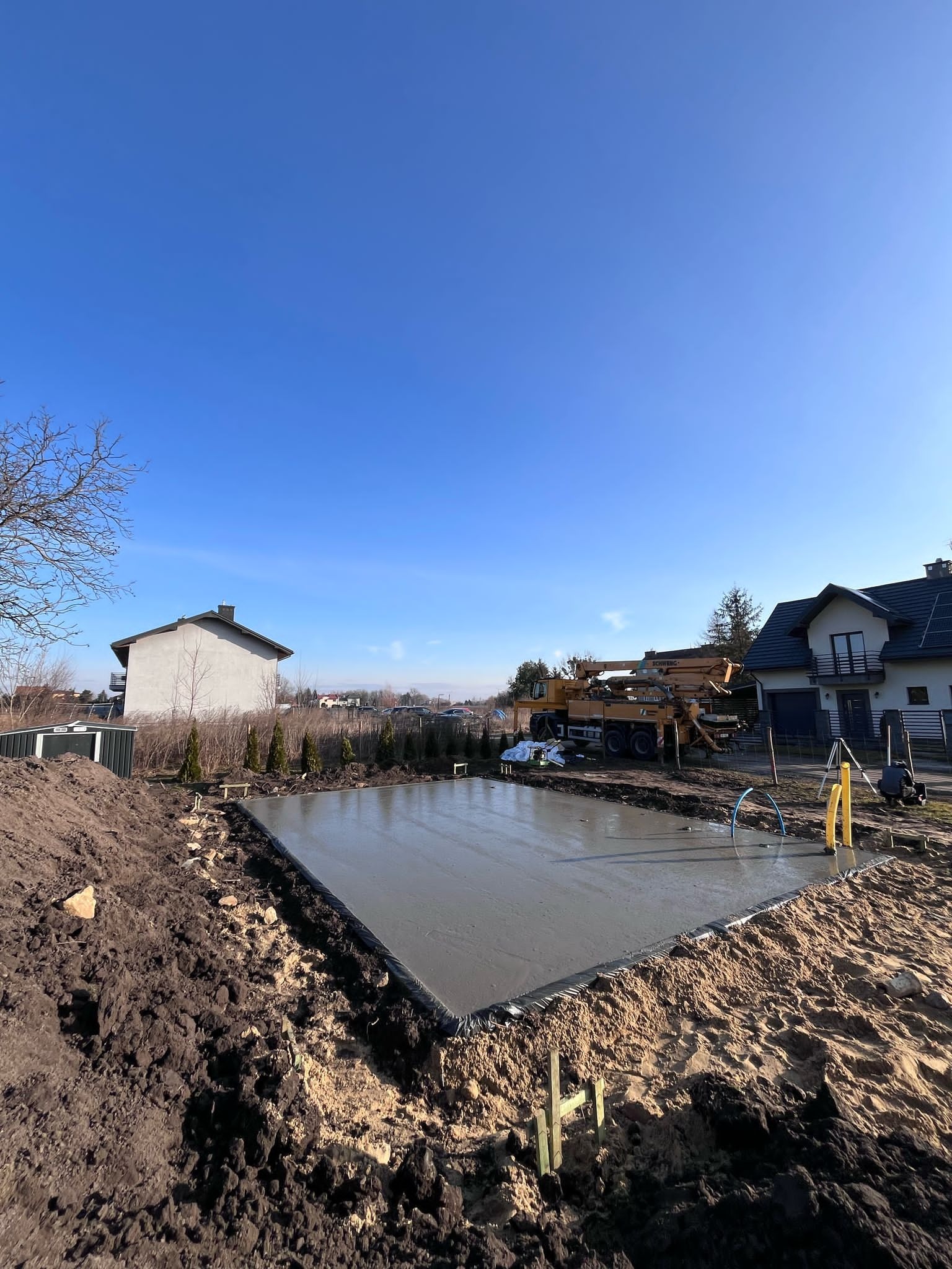 Świeżo wylany, gładki fundament pod budowę domu, otoczony ziemią, z widoczną pompą do betonu Schwing i domami w tle pod błękitnym niebem.