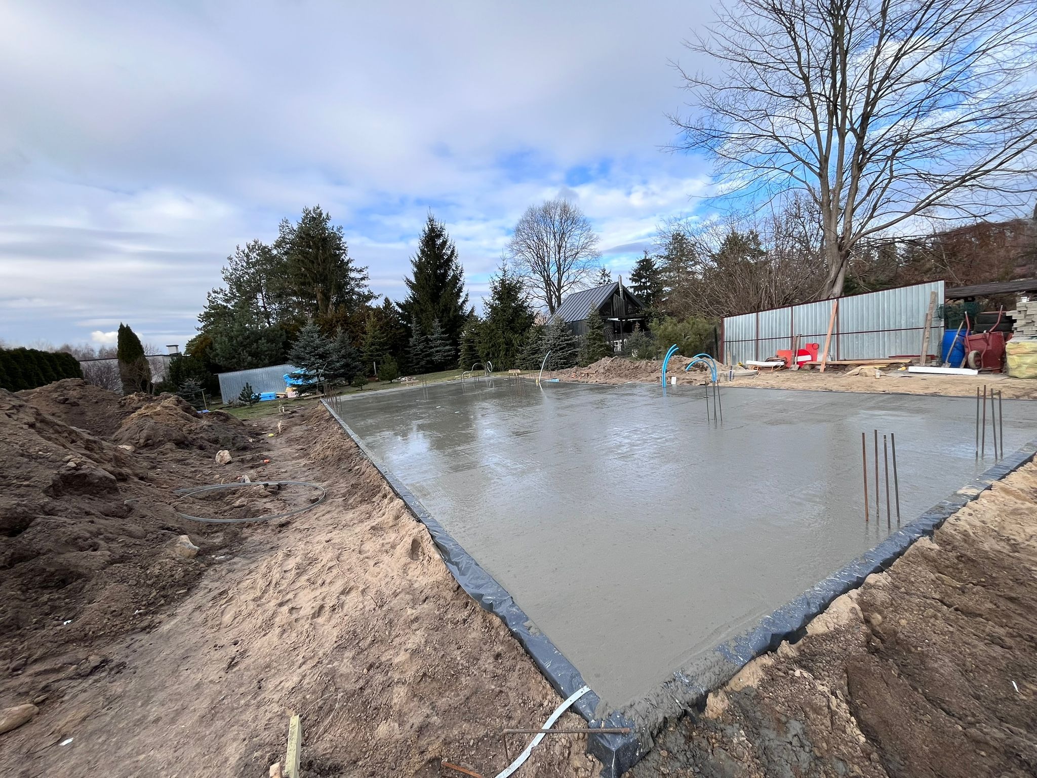 Świeżo wylany fundament płyty betonowej z wystającymi prętami zbrojeniowymi, zabezpieczony folią, otoczony ziemią, na tle drzew i pochmurnego nieba.