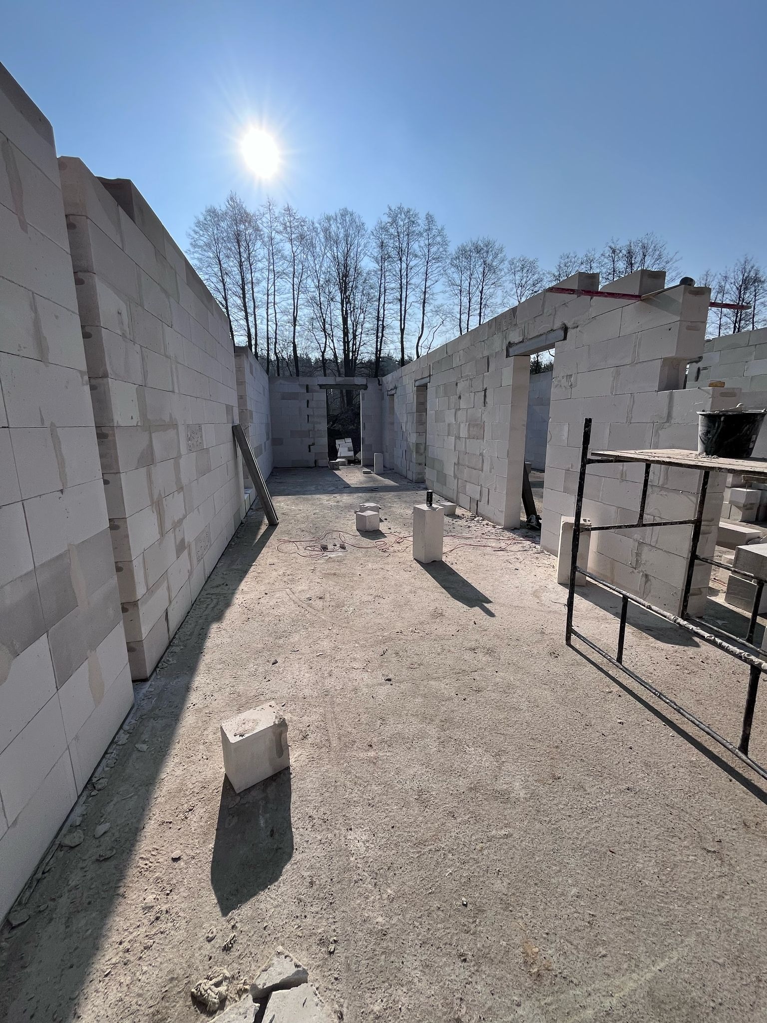 Wnętrze surowego domu z białych bloczków, widoczne otwory okienne i drzwiowe, w tle drzewa i błękitne niebo z mocnym słońcem.