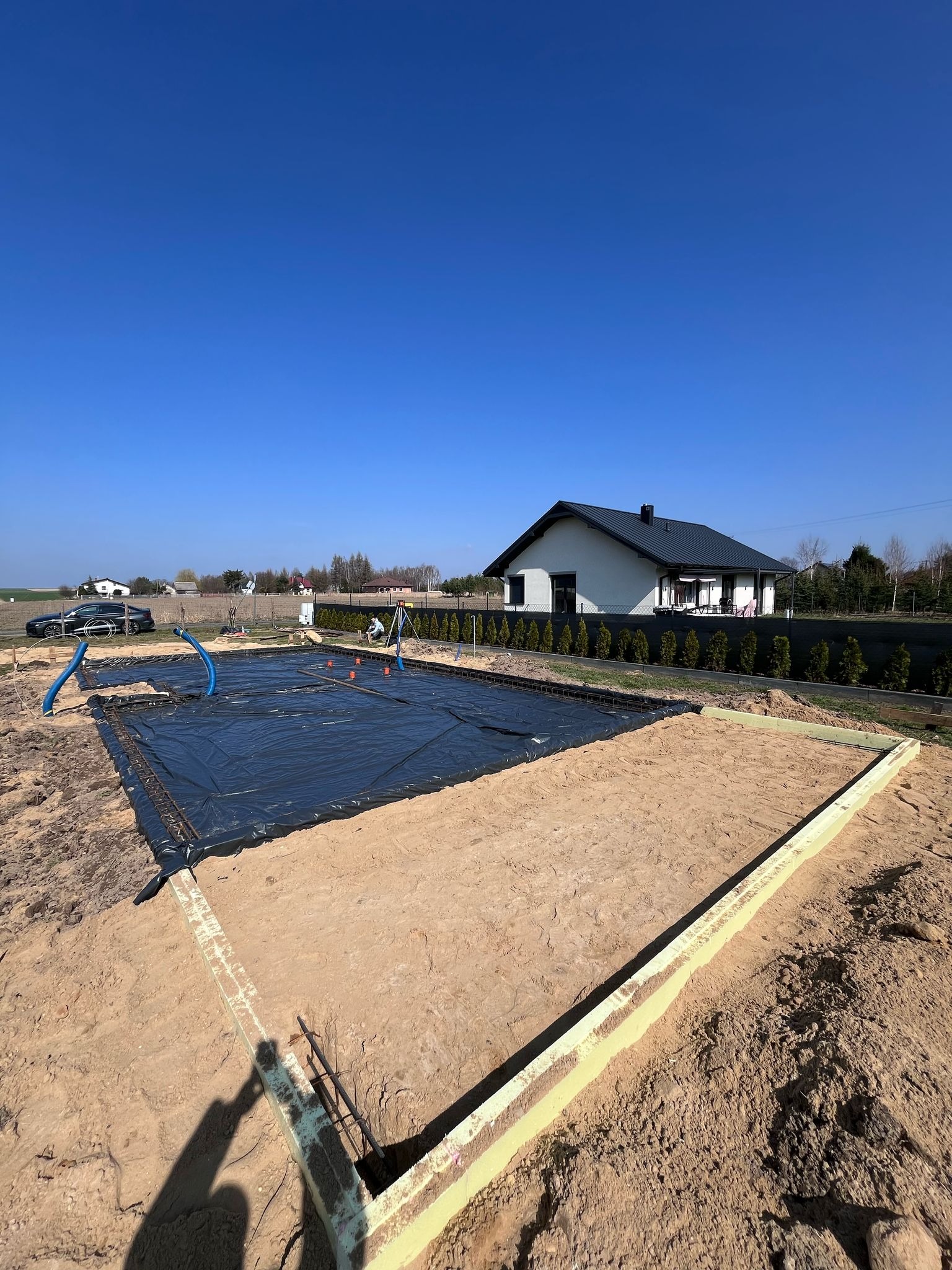 Przygotowanie terenu pod fundament: wykop z piaskiem, czarna folia izolacyjna, szalunki z izolacją termiczną, zbrojenie. W tle dom jednorodzinny i błękitne niebo.