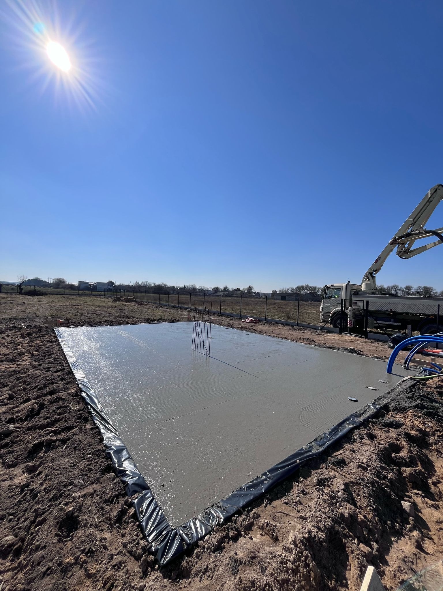 Świeżo wylany fundament pod budynek, zabezpieczony czarną folią, z wystającymi prętami zbrojeniowymi; na tle błękitnego nieba widoczny betonowóz z pompą.