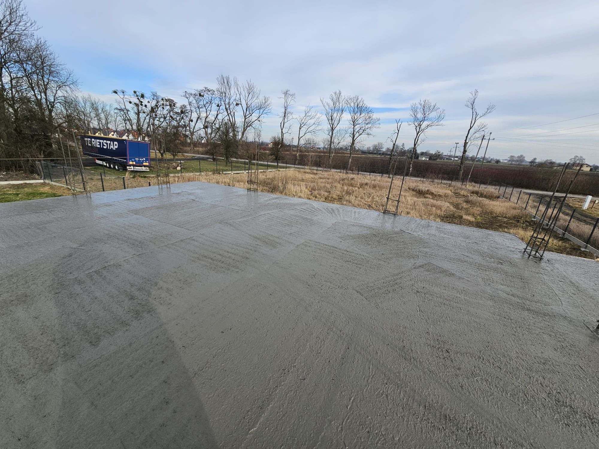 Świeżo wylana płyta fundamentowa z widocznymi śladami poziomowania, na tle zimowego krajobrazu z drzewami bez liści i ciężarówką w oddali.