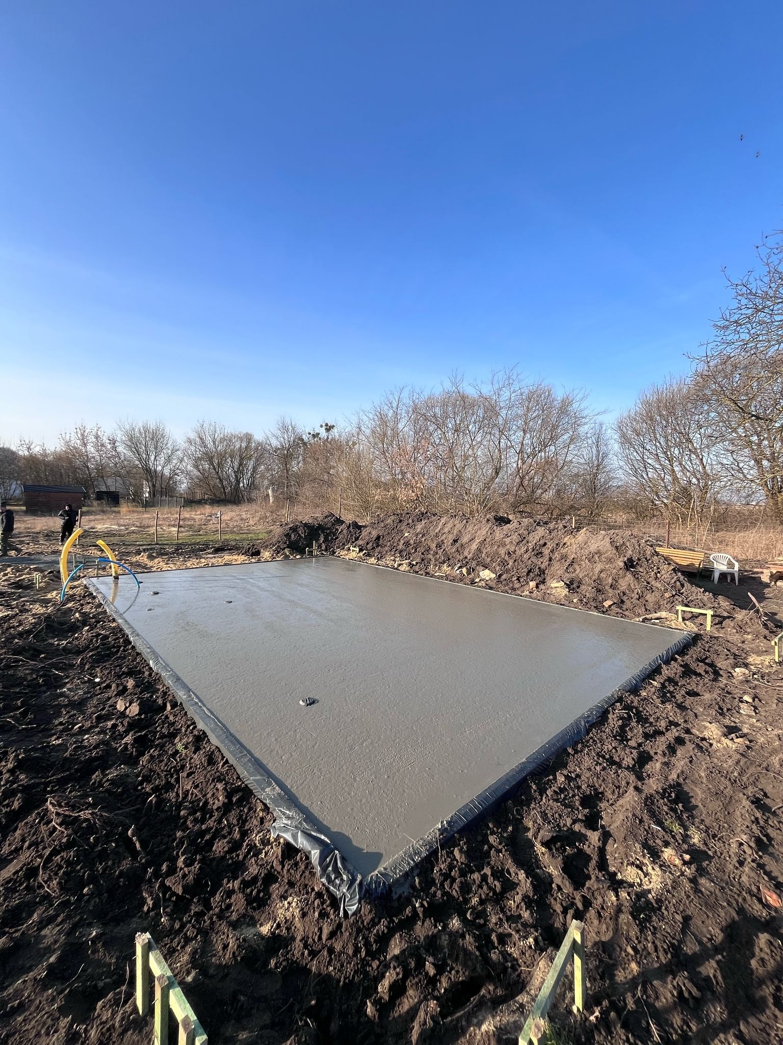 Świeżo wylany fundament pod budowę domu jednorodzinnego, zabezpieczony folią, widoczny żółty wąż do pompy betonu, w tle drzewa bezlistne i fragment zabudowań.