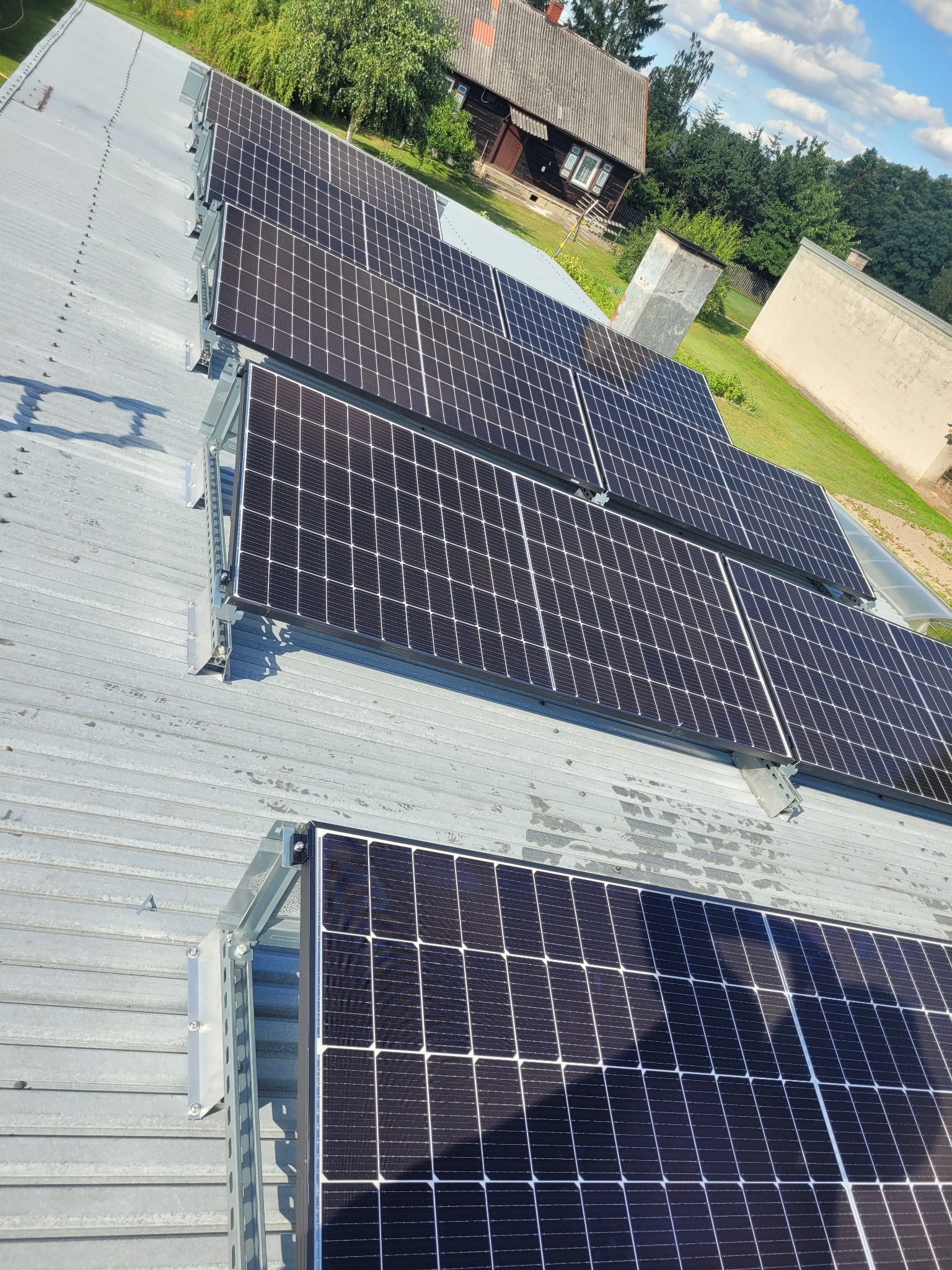 Rząd paneli słonecznych zamontowanych na metalowym dachu, widok z góry pod kątem, w tle budynki i drzewa.