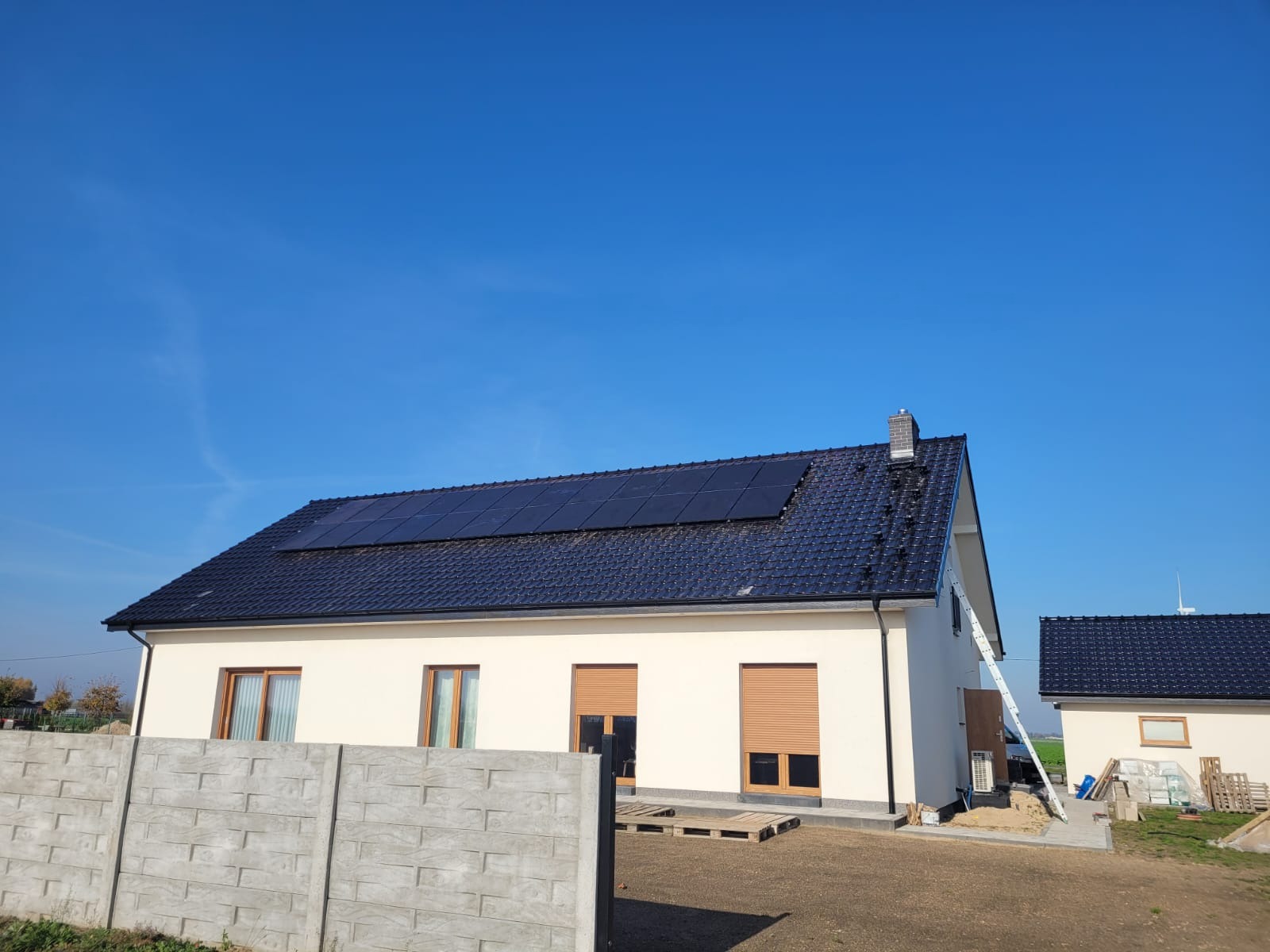 Dom jednorodzinny z panelami słonecznymi na czarnym, błyszczącym dachu, drabina oparta o ścianę, błękitne niebo.