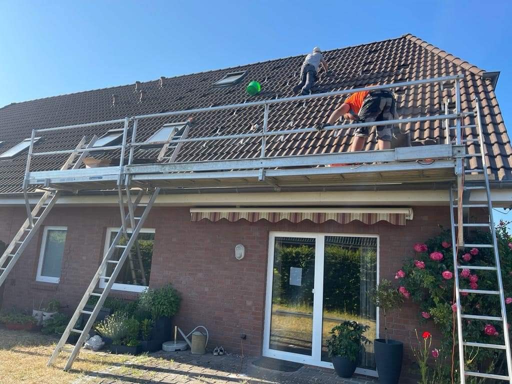 Montaż paneli na dachu domu jednorodzinnego z rusztowania; widoczne dwie osoby pracujące na dachu, narzędzia i elementy konstrukcyjne, zielona piłka na dachu.