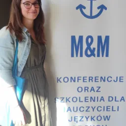 Uśmiechnięta kobieta w okularach i jeansowej kurtce stoi obok baneru z napisem 'M&M Konferencje oraz szkolenia dla nauczycieli języków obcych' i symbolem kotwicy.