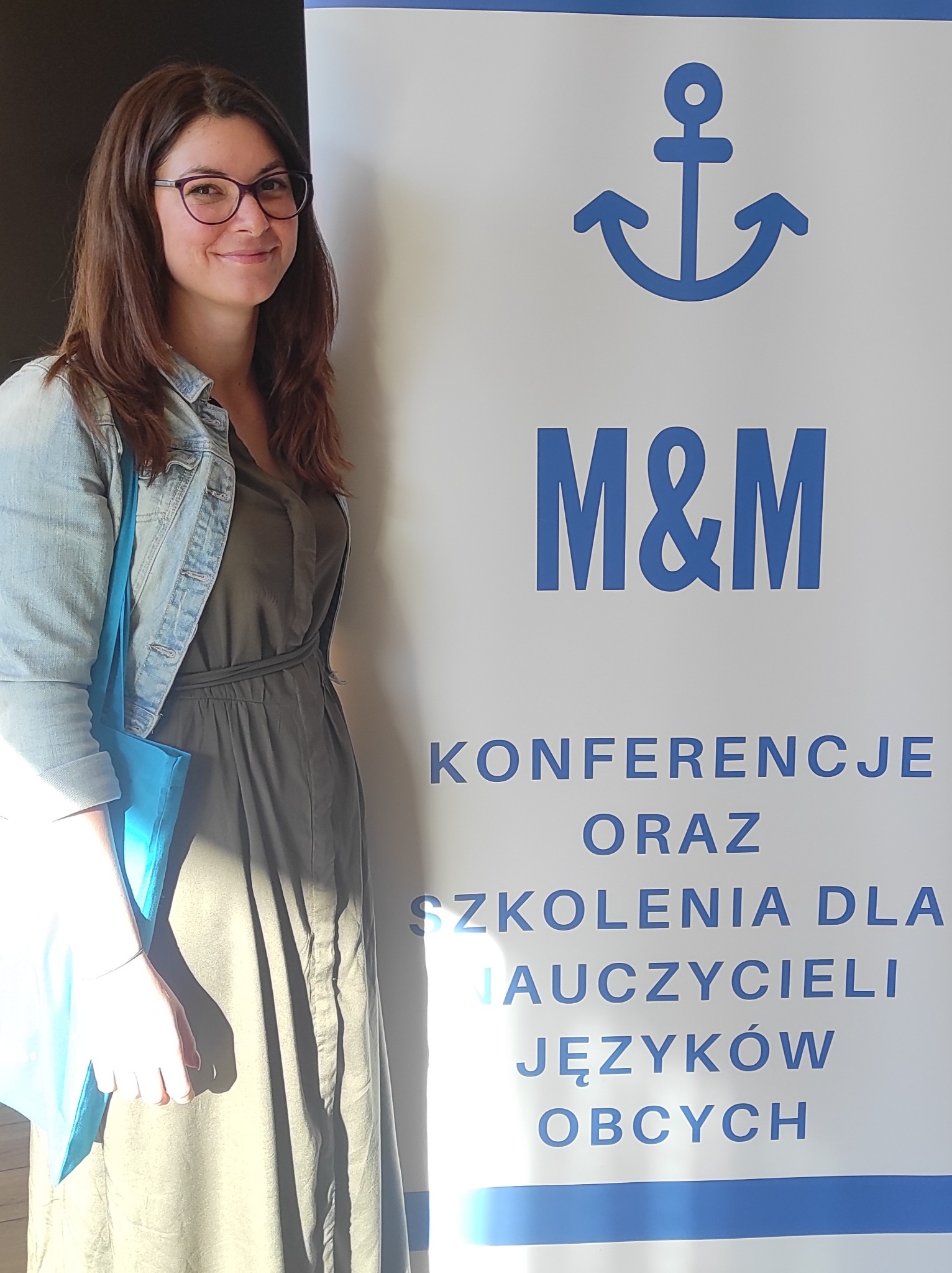 Uśmiechnięta kobieta w okularach i jeansowej kurtce stoi obok baneru z napisem 'M&M Konferencje oraz szkolenia dla nauczycieli języków obcych' i symbolem kotwicy.