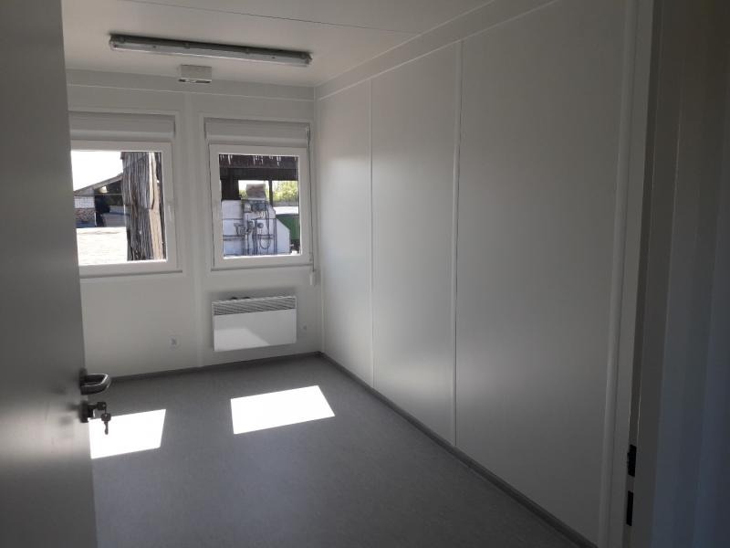 Wnętrze modułowego biura z dwoma oknami, grzejnikiem elektrycznym i szarą podłogą, widok z otwartych drzwi.