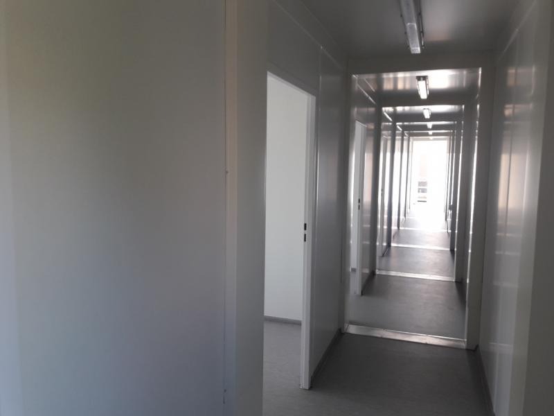 Wnętrze korytarza kontenera biurowego z szarymi ścianami, podłogą i oświetleniem fluorescencyjnym, widok w głąb z otwartymi drzwiami po lewej stronie.