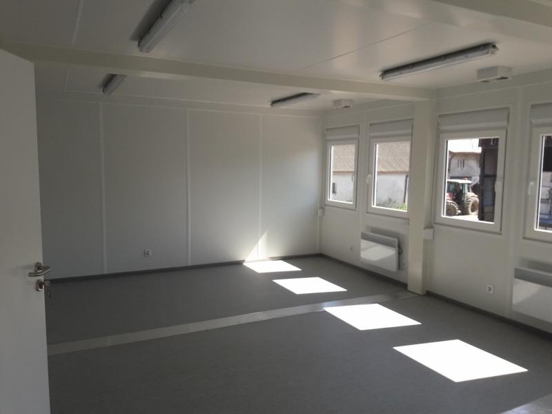 Wnętrze pustego, modułowego biura z trzema oknami, przez które widać traktor na zewnątrz, białe ściany z widocznymi łączeniami paneli, szara podłoga i białe grzejniki pod oknami.