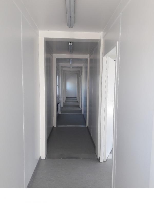 Wnętrze długiego, pustego, modułowego korytarza z szarą podłogą i białymi ścianami, oświetlonego przez lampy sufitowe
