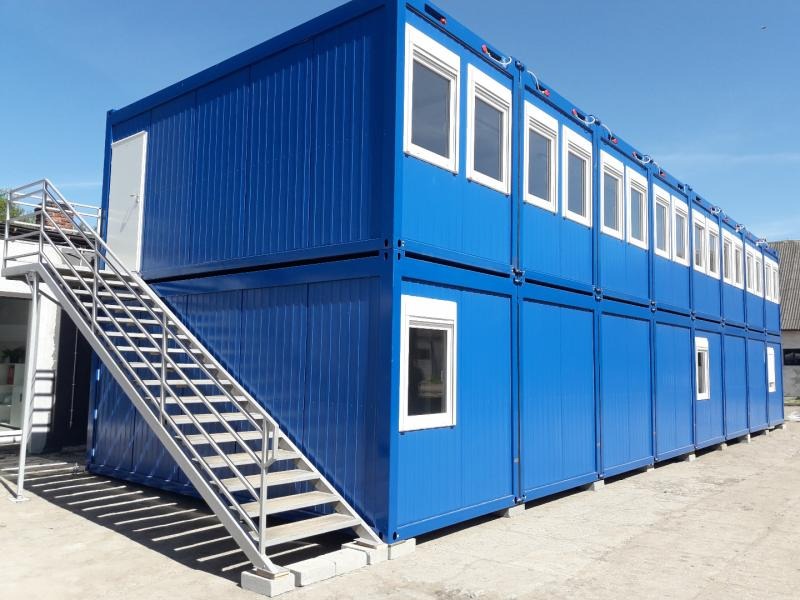 Dwukondygnacyjny, modułowy budynek biurowy z kontenerów w kolorze niebieskim, z zewnętrznymi schodami i białymi oknami, na placu budowy.