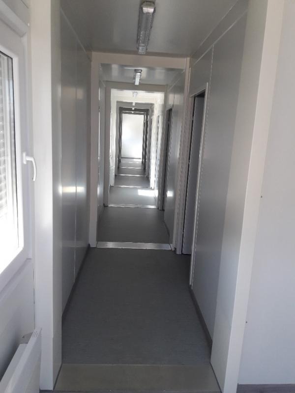 Długi, wąski korytarz wewnątrz modułowego budynku z szarym linoleum na podłodze i białymi ścianami, oświetlony lampami jarzeniowymi na suficie.