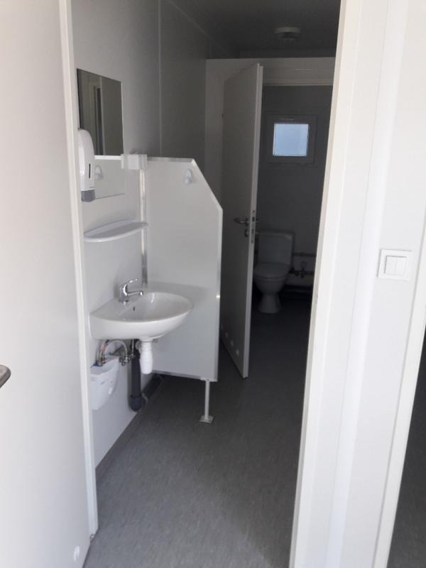 Wnętrze przenośnej toalety z umywalką, dozownikiem mydła i toaletą w tle, widok z zewnątrz przez otwarte drzwi.