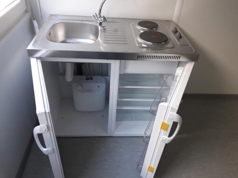 Kompaktowa, mobilna kuchnia z nierdzewną umywalką, dwupalnikową płytą elektryczną i lodówką z otwartymi drzwiami, widoczny biały zbiornik na wodę pod zlewem.