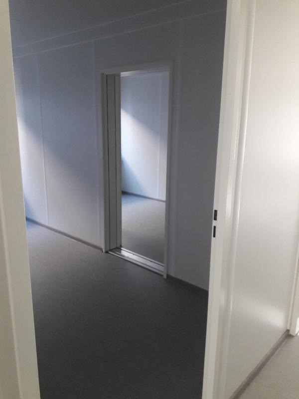 Wnętrze modułowego pomieszczenia biurowego z szarą podłogą i przesuwnymi drzwiami, widok przez otwarte drzwi.