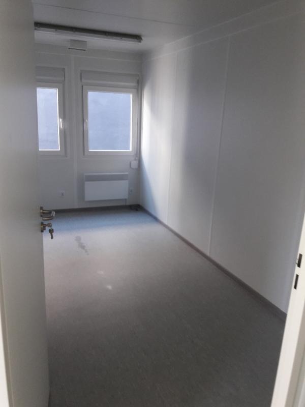 Wnętrze pustego, jasnego kontenera biurowego z dwoma oknami, grzejnikiem i szarą podłogą, widok zza otwartych drzwi z kluczem w zamku.