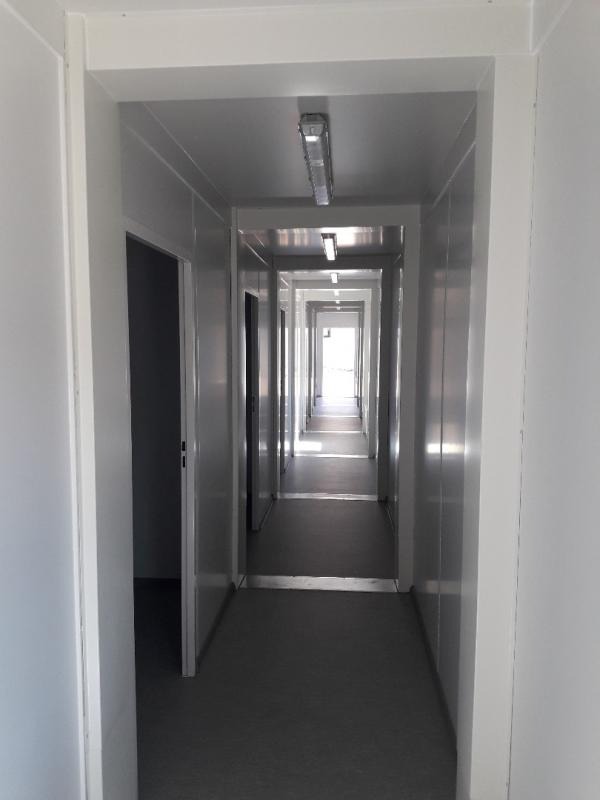 Długi, pusty korytarz wewnątrz kontenera biurowego z widocznymi lampami sufitowymi i otwartymi drzwiami po lewej stronie.