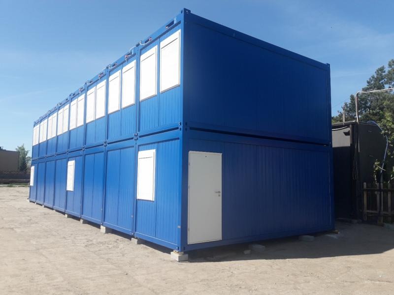 Dwu-kondygnacyjny, niebieski kontener biurowy z białymi oknami i drzwiami, ustawiony na betonowych blokach na placu budowy.